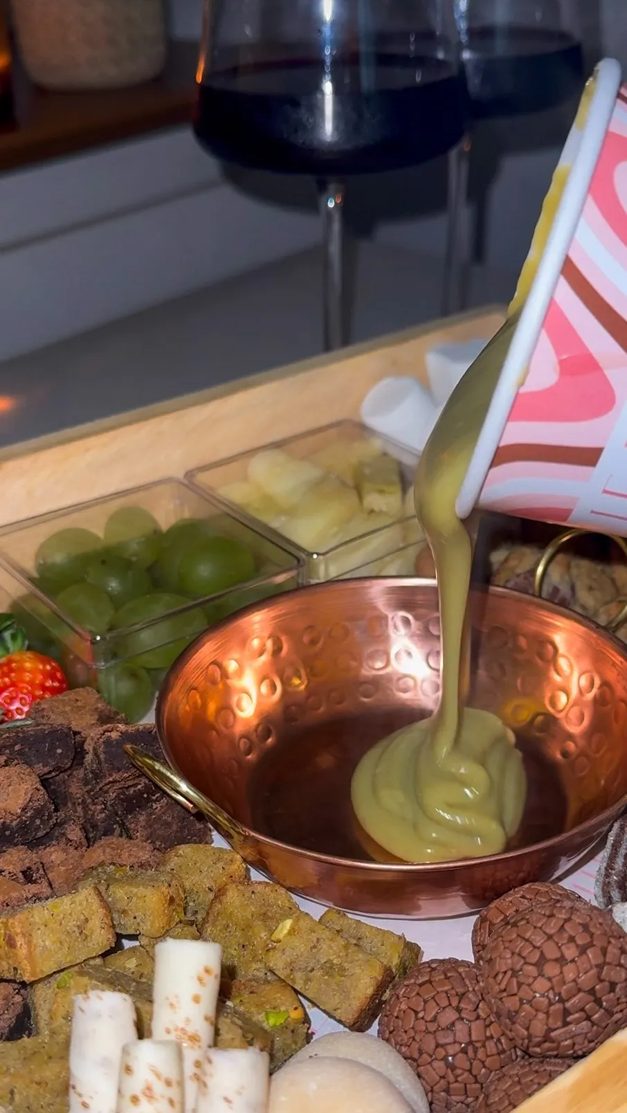 Creme claro sendo despejado de um pote numa panelinha de cobre martelado, cercada por brownies, fatias de bolo verde, brigadeiros e uvas numa tábua de madeira.