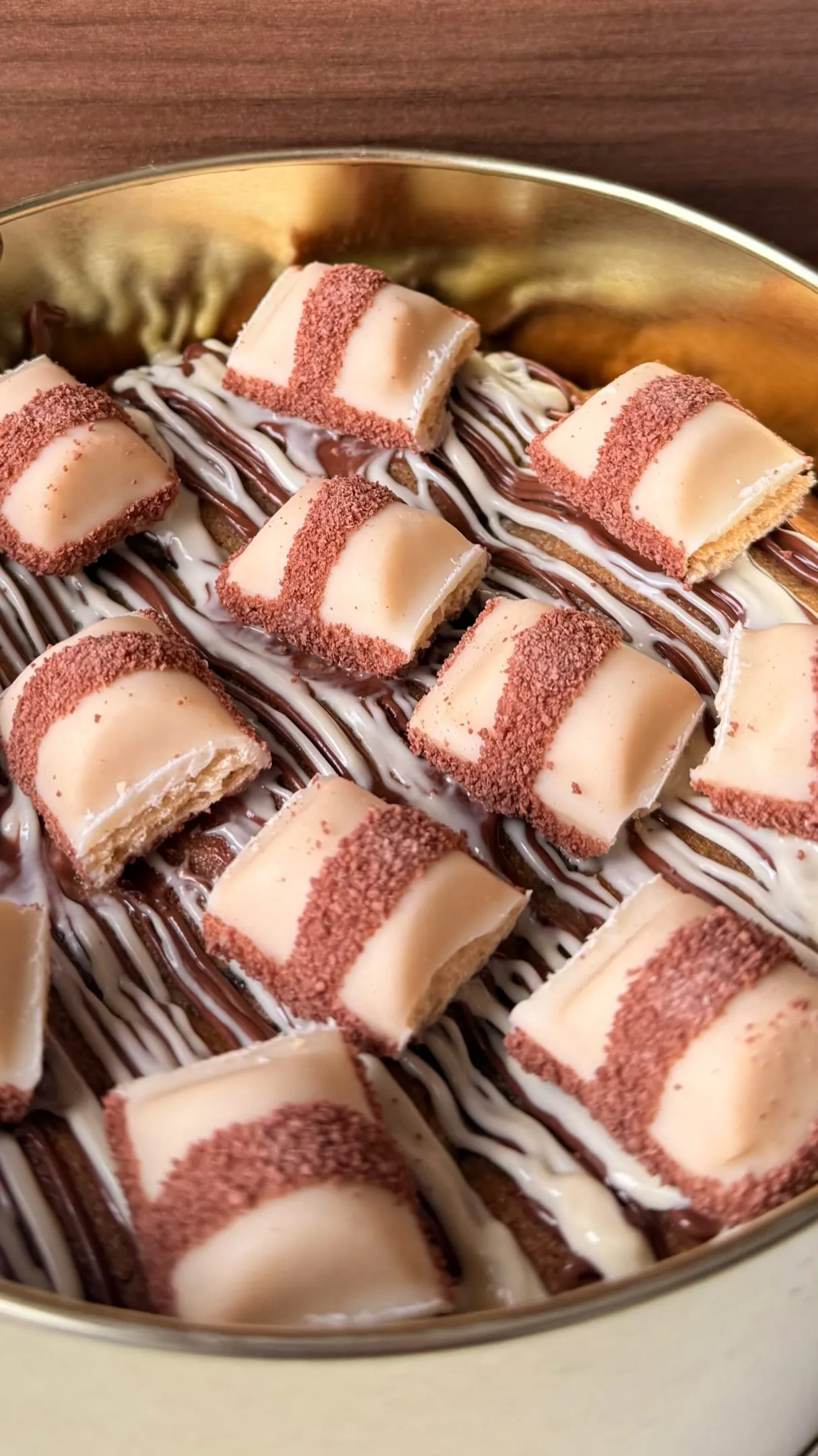 Lata dourada forrada com fios de chocolate ao leite e branco, coberta de quadradinhos de doce claro polvilhados de rosa.