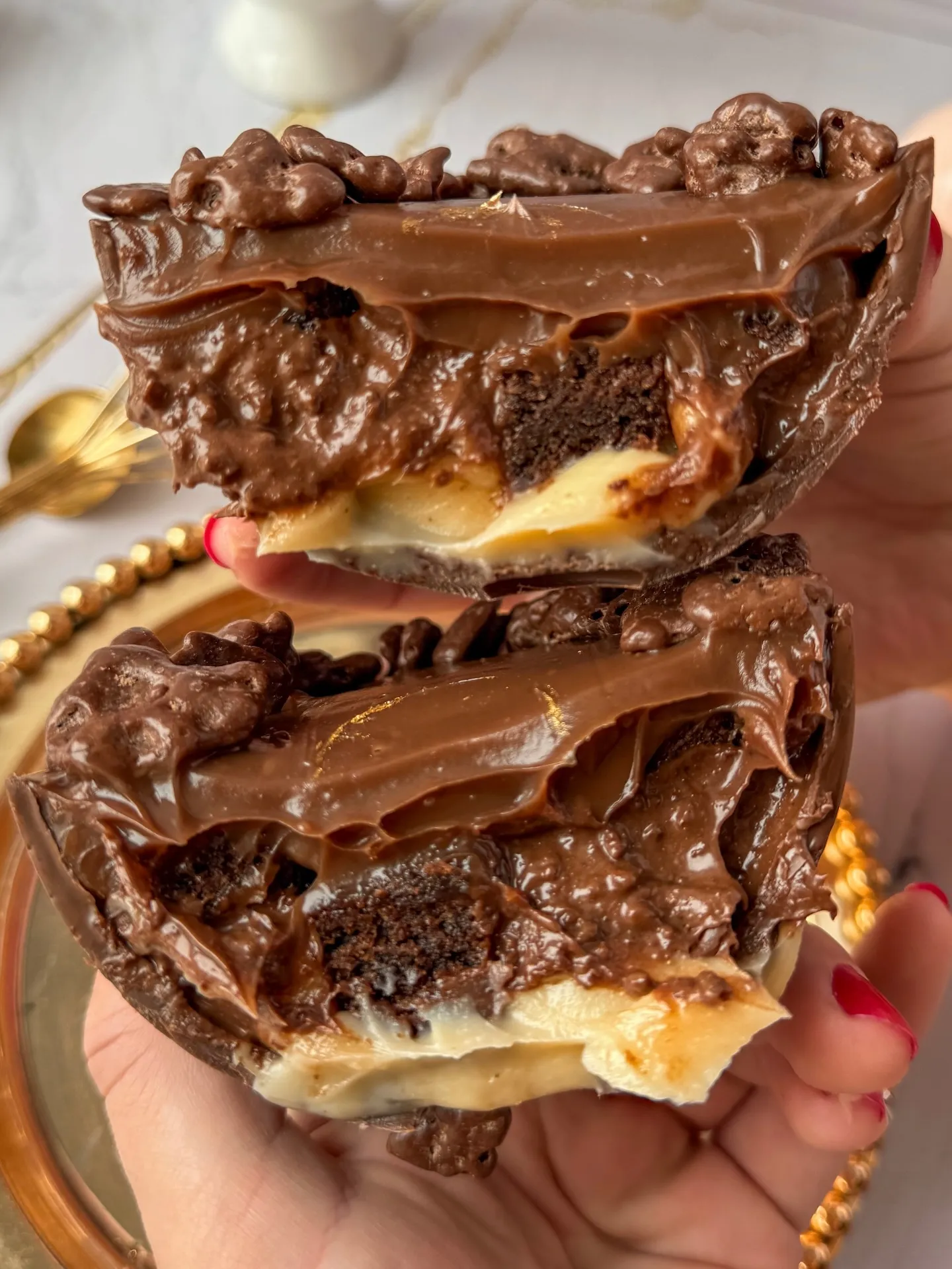Duas metades de ovo de chocolate empilhadas numa mão, mostrando as camadas por dentro: bolo de chocolate, creme claro e cobertura crocante.