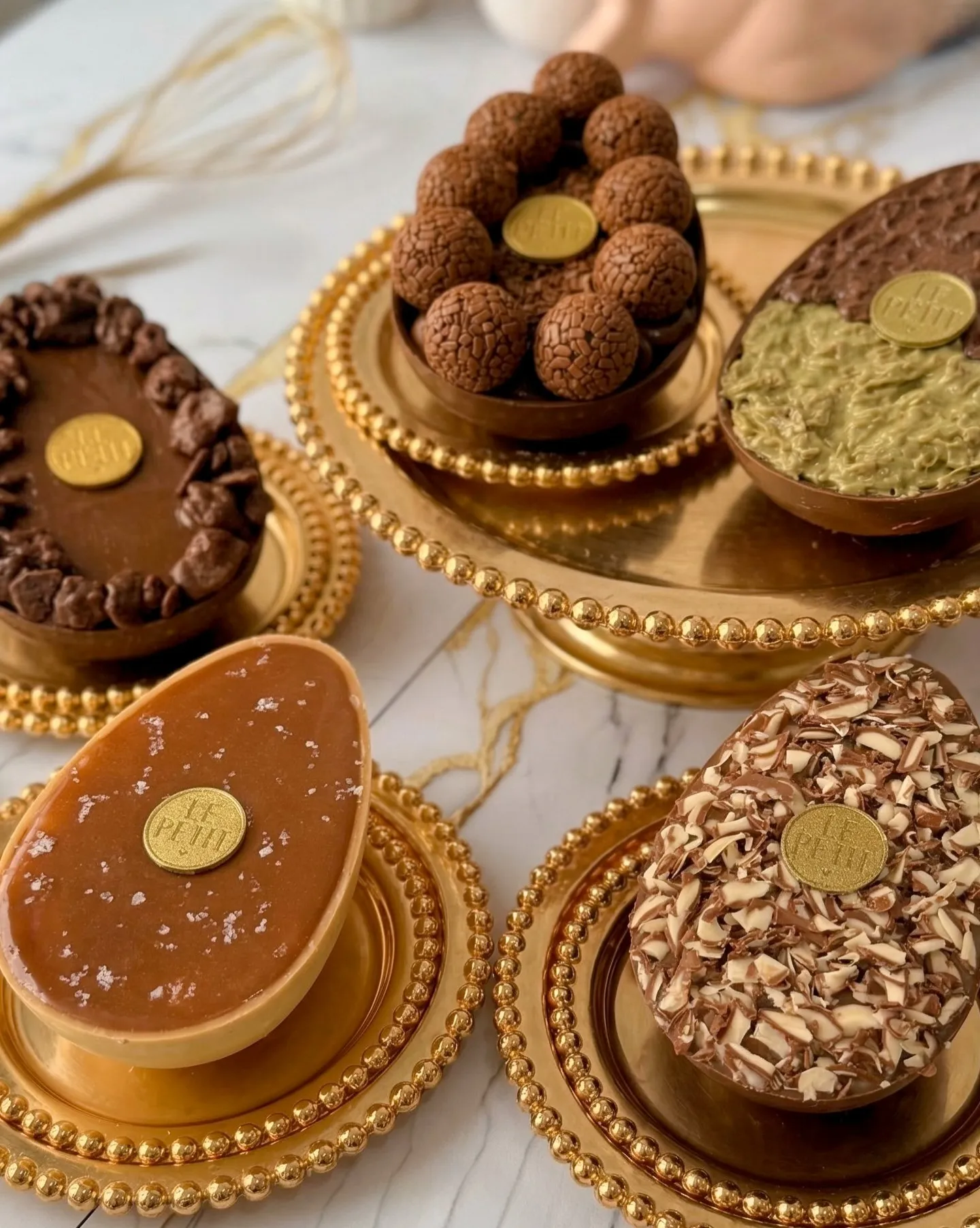 Cinco ovos de chocolate sobre pratos e bandejas dourados de borda em contas, cada um com um selo dourado Le Petit: um de brigadeiros, um de pistache, um de caramelo salgado e dois de chocolate.