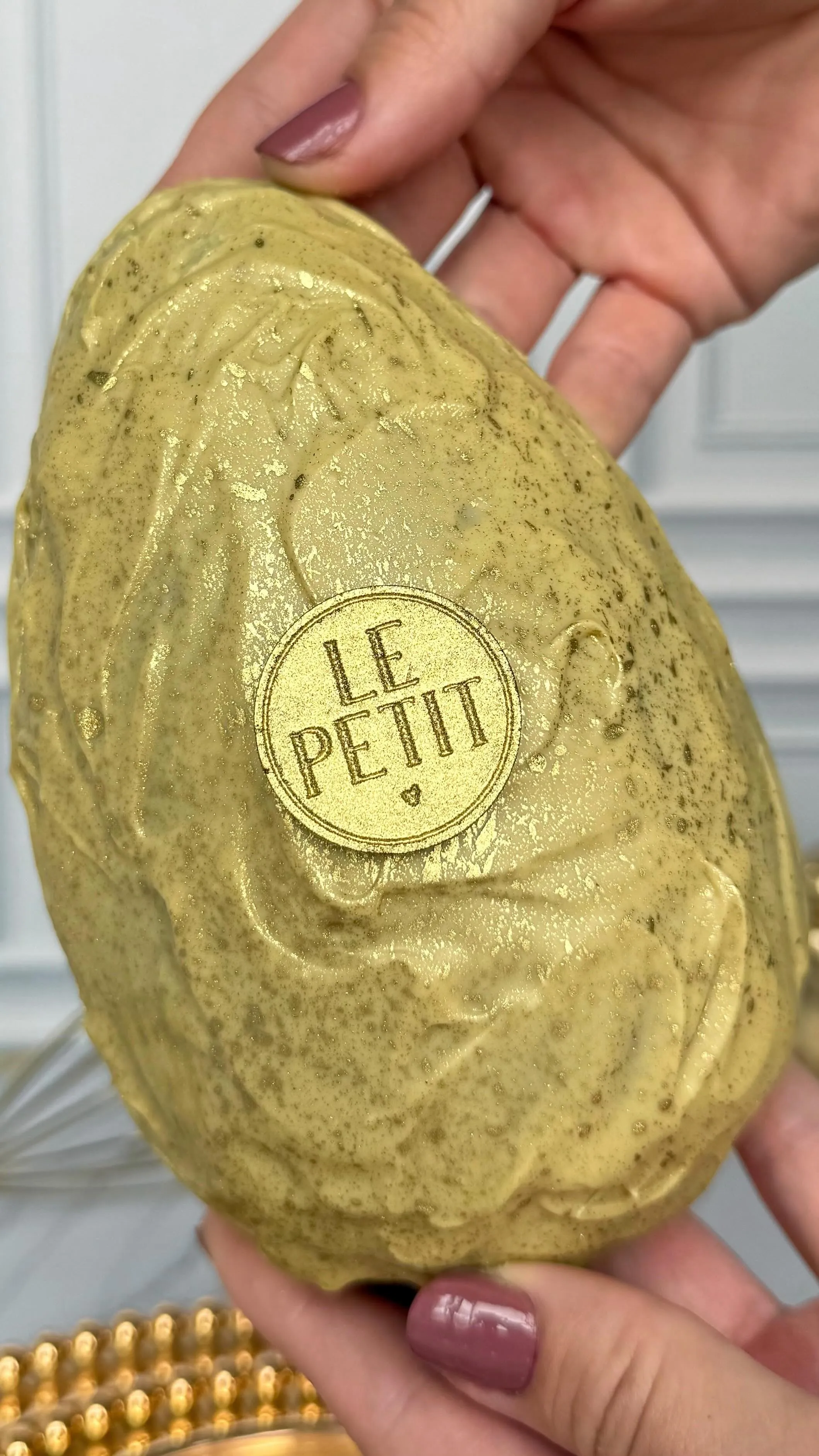 Ovo coberto por creme de pistache salpicado de dourado, com o selo Le Petit no centro, segurado por duas mãos.