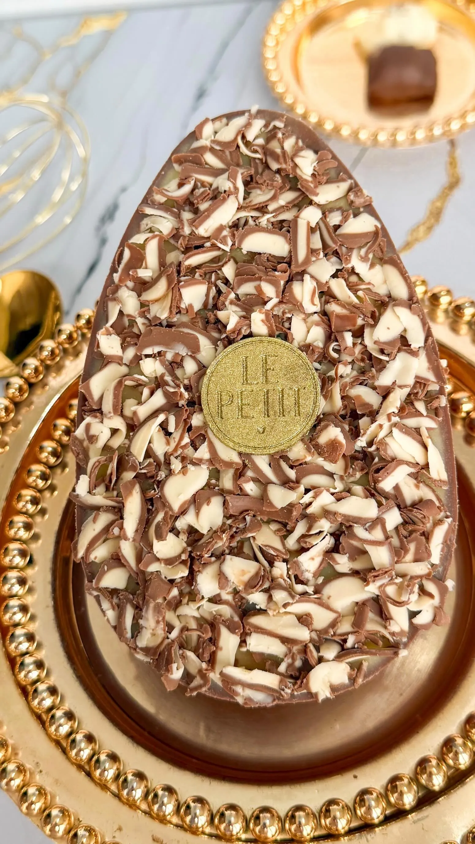 Ovo coberto de raspas de chocolate ao leite e branco, com o selo dourado Le Petit, sobre um prato dourado de borda em contas.
